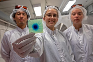 Three scientists in cleanroom attire—Kei Nakamura, Lieselotte Obst-Huebl, and Anya Jewell -- show a radiochromic film that has been hit by the proton beam at BELLA’s iP2. 