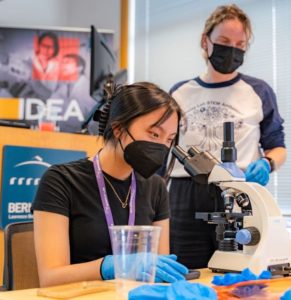 A Berkeley Lab STEM Ambassador looks on as the young woman she is mentoring uses a microscope