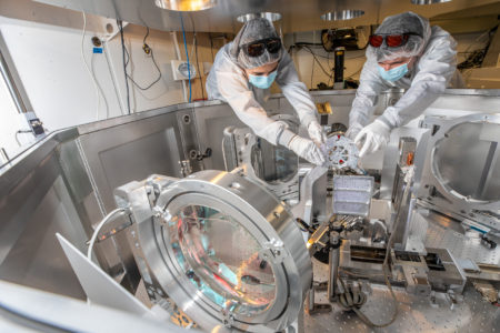 Two researchers in cleanroom attire work on a laser apparatus