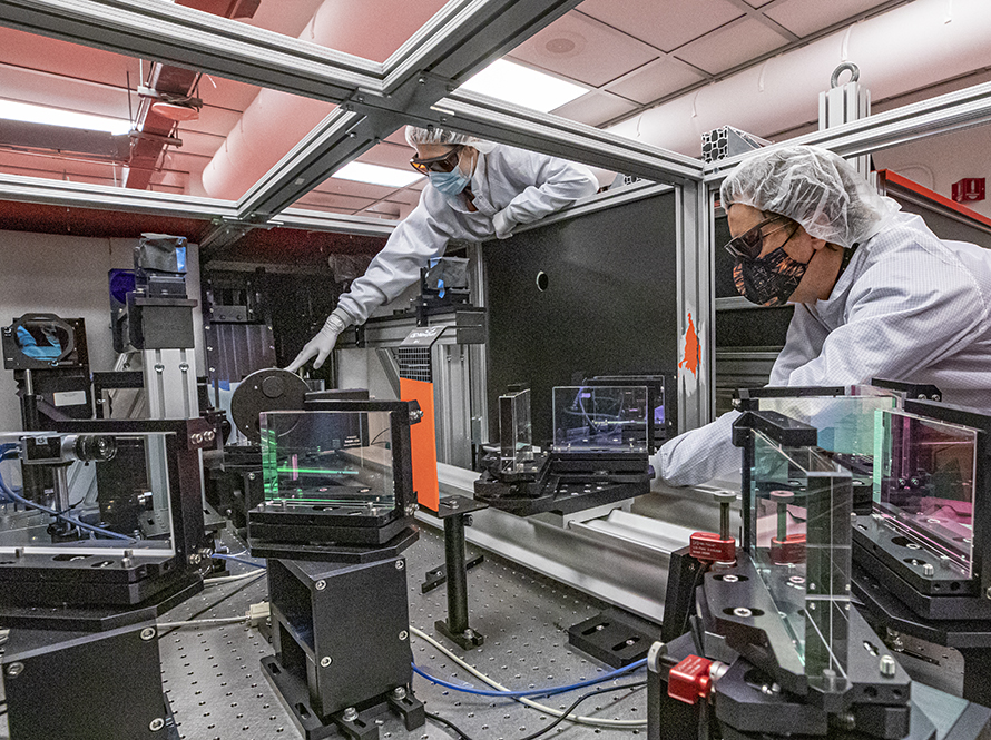 Researchers Marlene Turner and Tony Gonsalves, gowned and goggled for a laser area, work at an optical table on a “staging” experiment