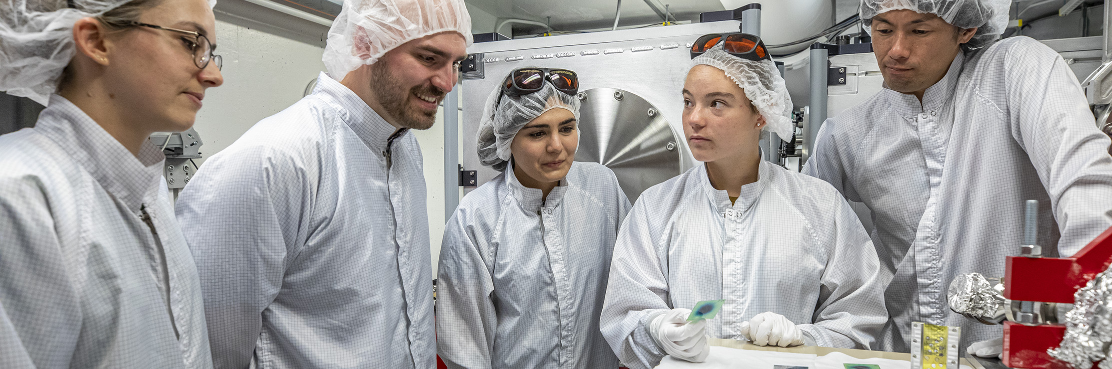 Several researchers in cleanroom attire discuss beam images in a laser lab