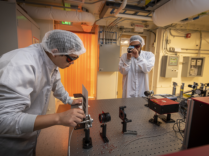 Researchers gowned and goggled for a laser area, work at an optical table on a beam combining experiment