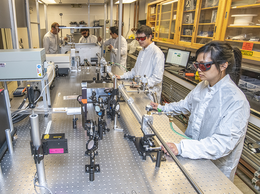 Five researchers, gowned and goggled for a laser area, work at a long optical table