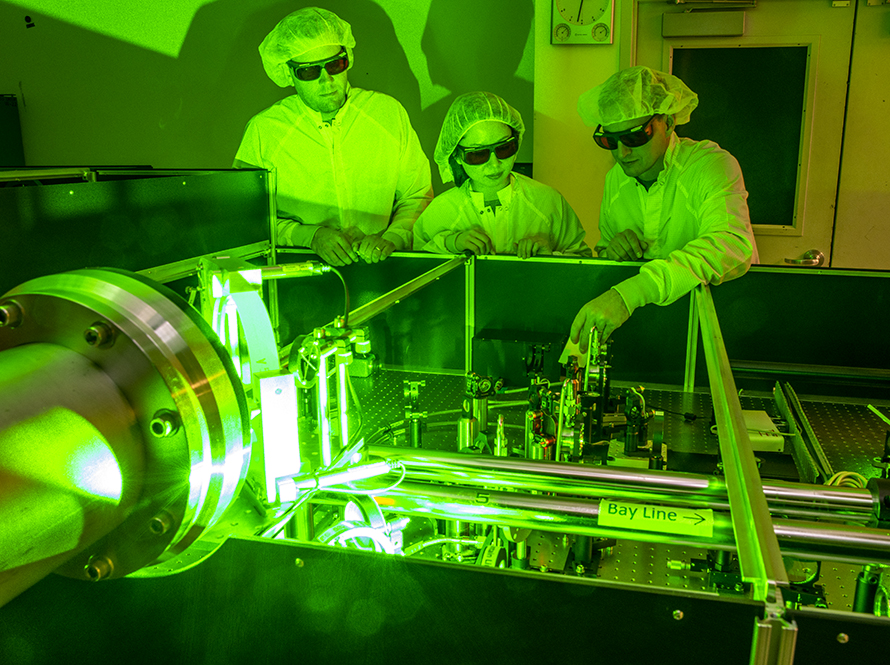 Three researchers, gowned and goggled for a laser area, bathed in green light, work on an experiment