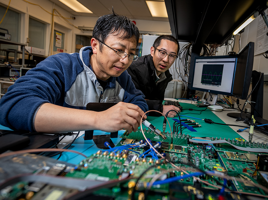 Two researchers probe a circuit board on a test bench