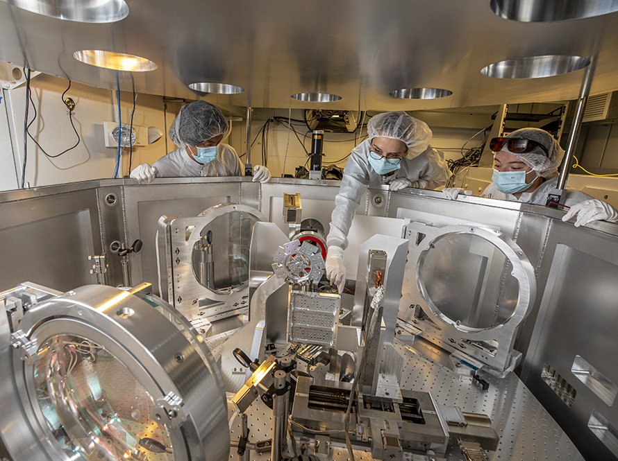 Three researchers wearing cleanroom gowns, white gloves, and hairnets work in a laser area