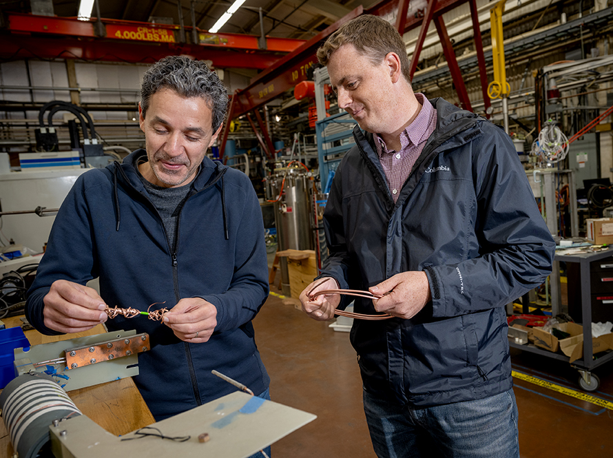 Two scientists in a lab/shop area examine superconducting wire